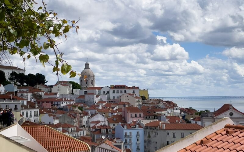 Telhados vermelhos e uma cúpula sobre Alfama, com o rio Tejo ao fundo.